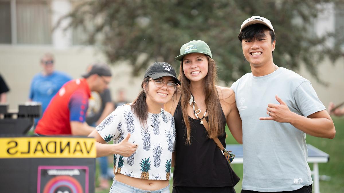 Smiling dark haired young man with baseball hat on head gives 'hang loose' hand gesture on grassy lawn with two smiling dark-haired young women in baseball caps.